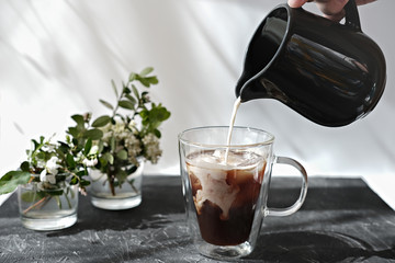 hand holding milker and pouring milk into a coffee mug. cold coffee with ice cubes and flowers on a grey table. morning breakfast time. relax concept.