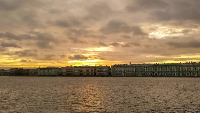 Cityscape of Saint Petersburg at the sunrise. The sun rises directly above the buildings on the banks of the Neva River. Small waves on the Neva river. Dark cumulus clouds moves across the sky.
