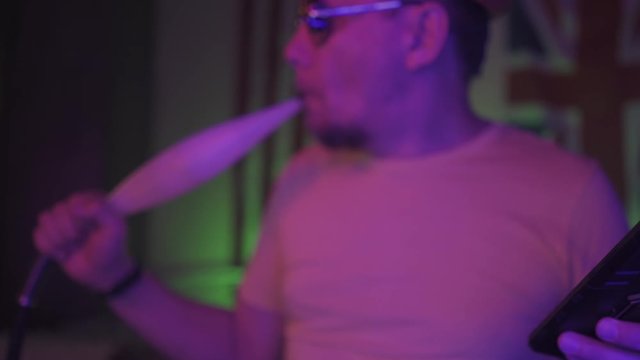 Man looking at the screen of a smartphone while smoking hookah, sitting in the room with lit neon blue and red lights with USA and UK flags on the wall. 