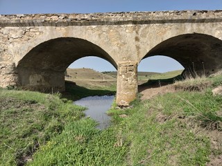 Old Soviet bridge made of stone. A small stream in the middle of green grass and flowers. Blue sky.