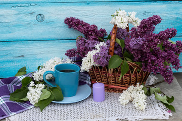 On a white knitted napkin is a cup of tea. Nearby is a bouquet of white and purple lilacs in a basket and a candle.