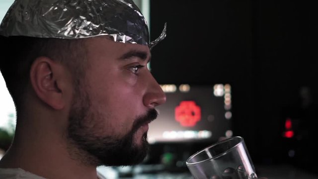 Man in tin foil hat watching white TV at home and drinking