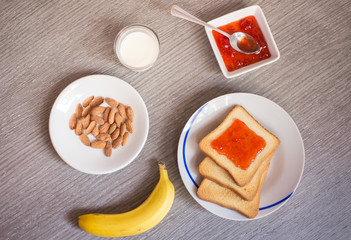 Top view of a meal made with a toast with strawberry marmalade, milk, a banana and a plate of almonds over the table