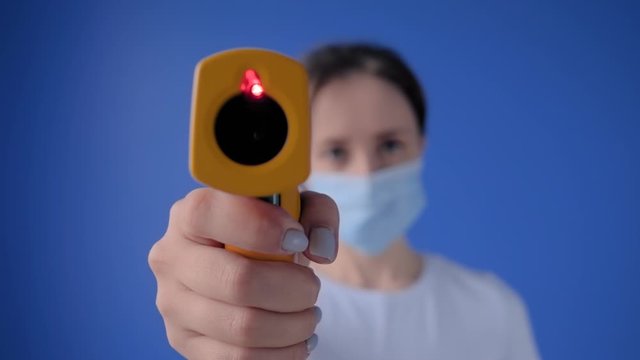Slow motion: woman in medical face mask holding yellow pyrometer to measure temperature toward camera - close up view, selective focus. Healthcare, measurement, disease, infection, coronavirus concept