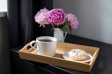 Three red peonies in a white porcelain jug, a cup of tea, a donut on a plate and a napkin for breakfast on a black table.