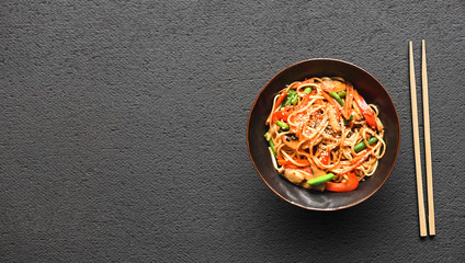 Chopsticks and bowl with wok noodles and vegetables on a black table background with a copy space.