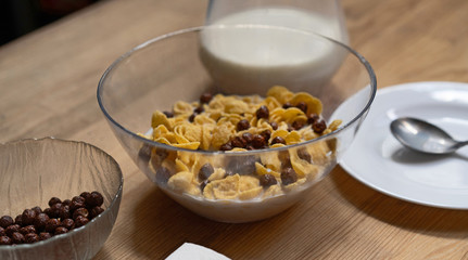 Cornflakes with chocolate balls and milk in a glass bowl on a wooden table with a jug of milk and a plate with a spoon on it. Healthy food concept. Quick breakfast concept