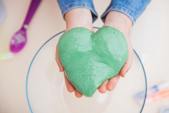 Chilld's hands holding green slime in heart shape. Dye how to make fluffy toy. Favourite handmade toy slime.