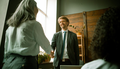 Business handshake of young businessman and gray-haired businesswoman looking at each other standing at office desk after team brainstorming in office. Shot from below