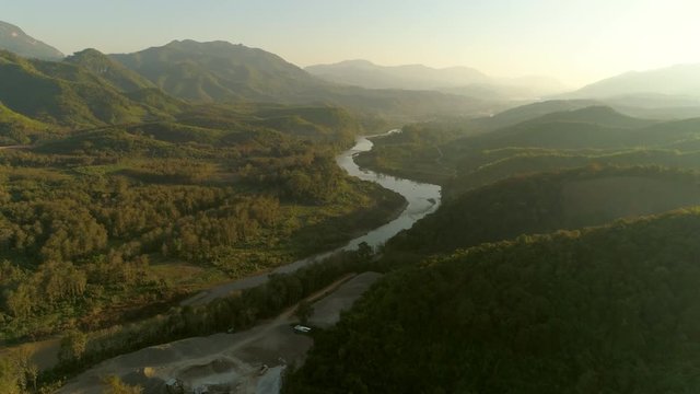Aerial shot of river amidst green forest against sky during sunrise, drone flying backward over landscape - Tat Sae Waterfalls, Laos