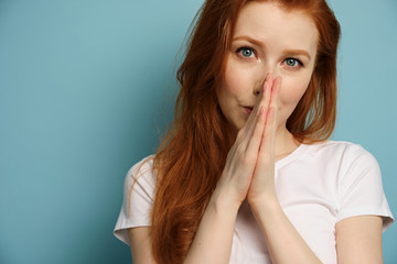 A red-haired girl in a white T-shirt stands on a blue background, pressing her palms to her face.