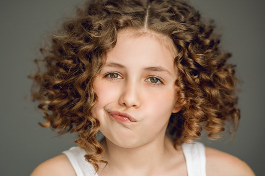 Close-up portrait. A teenage girl with curly hair curled her lips into a tube. The vzgyad directs directly into the frame. Gray background. Emotions thoughtfulness, surprise, smile, straight teeth.
