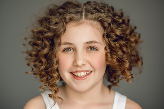 Close-up portrait. Teen girl with curly hair smiles beautifully. The vzgyad directs directly into the frame. Gray background. Emotions joy. Delight, smile, even teeth.