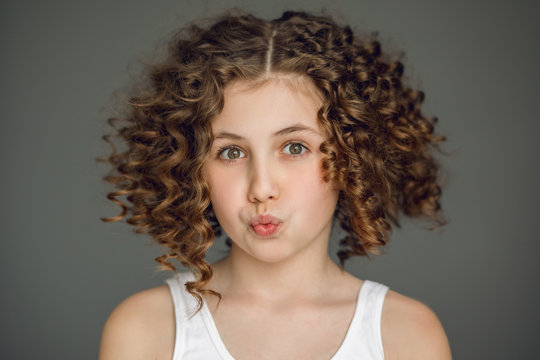 Close-up portrait. A teenage girl with curly hair curled her lips into a tube. The vzgyad directs directly into the frame. Gray background. Emotion surprise, joy, delight, smile, straight teeth.