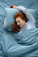 A red-haired girl in striped pajamas is lying in a blue bed stretching. Frame on top. 