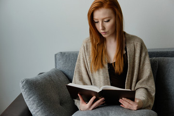 Red-haired girl in a warm cape sits on the sofa and carefully reads the black book, lowering her...