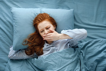 A red-haired girl in striped pajamas lies in a blue bed and yawns with her hand covering her mouth. Frame on top