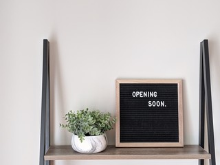 Coming Soon wood framed felt letter board with green plant in a pot on wood and metal bookshelf. Small and local business announcement teaser. Website and online news announcement coming soon. Updates