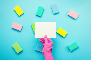 female hand in a pink rubber glove holds an empty card on a blue background with sponges. cleaning service concept. Copy space. Flat lay, Top view