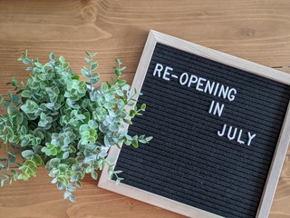 Re-opening/ reopening in July Announcement wooden framed letter board and potted plant on a wooden table. Layflat. Announcement. Small and local business news. Website/ blog and company updates.
