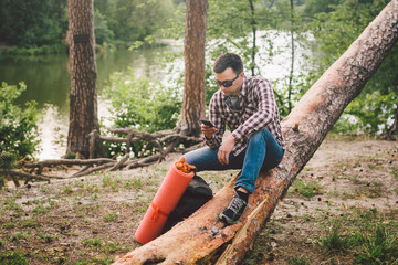 Naklejka na ściany i meble Young man sit on truck in forest and using mobile phone. man traveler sits on large fallen tree, holding phone in hand. theme hiking nature travel. Outdoor activities hiking. Traveling and adventure