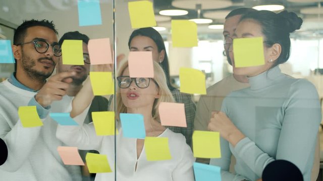 Colleagues multiracial team are working with sticky notes on glassboard busy with problem solving focused on creative activity. People and strategy concept.