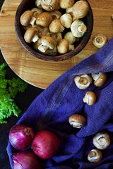 Wooden bowl of Portabellini mushrooms on a wooden board.  A blue cloth sits to the side with a few isolated mushrooms next to the bowl.  Red onions and a tuft of parsley sit to the right of the bowl