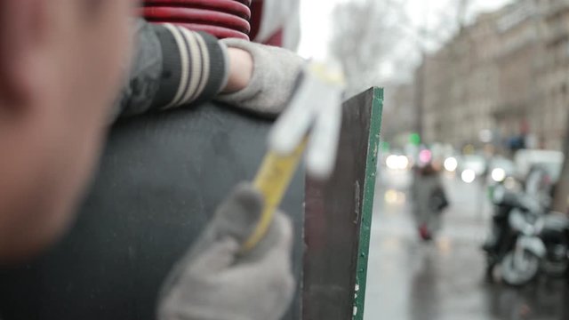 Men's Hands In Construction Gloves Hammer A Nail Into The Fence, There Is Dust. Close Up
