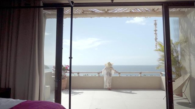 Young woman on vacation wearing hat, bikini and sheer dress stands on balcony just outside her bedroom taking in a breathtaking view during her vacation