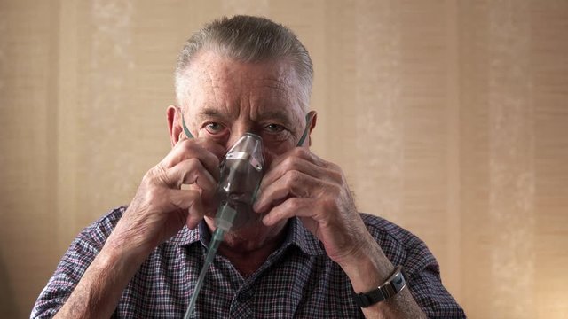 The old man putting on the breathing mask and makes few deep breaths. Portrait of the man with silver hair watching straight in the camera. The concept of medical treatment of respiratory diseases.