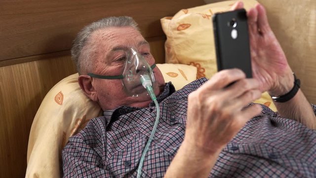 The Man In Breathing Mask Is Laying At The Bed And Looking At The Phone. Old Man Is Using Nebuliser And Pushing The Screen Of The Smartphone. Theme Of Medical Curation Of Various Lung Diseases.