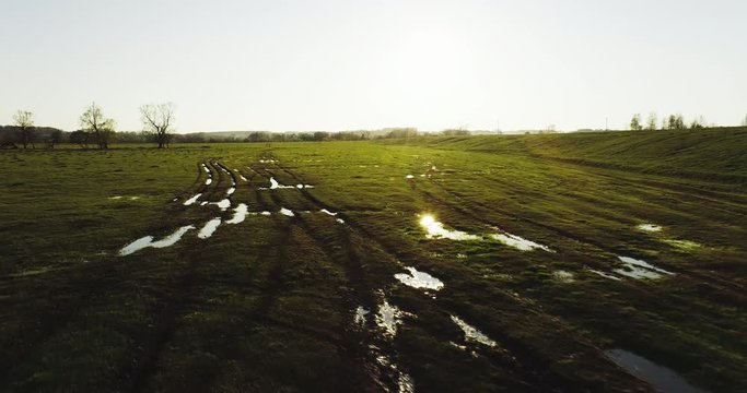 Dirt earthen roads with puddle with sun glare reflection in flat grass field near floodplain river. Wide drone shot flooded area at spring time cloudy day