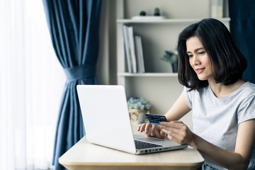 Young beautiful Asian woman holding credit card shopping online from home. Girl using laptop computer for buy items from internet during Covid19 pandemic at house. New normal modern purchase concept.