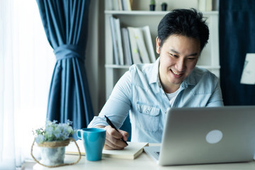 Asian businessman talking to colleague team in video call conference writing note on book with smile face. Man using computer laptop for digital online meeting network. Smart working from home concept