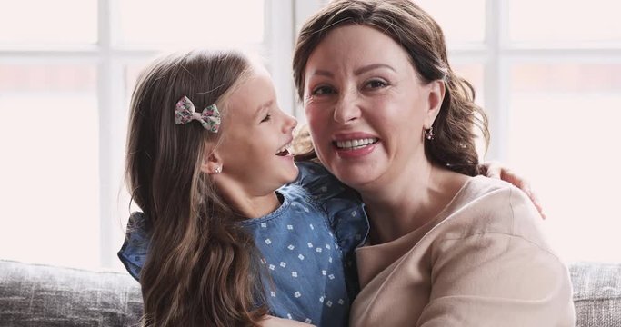 Affectionate middle aged smiling woman cuddling little preschool granddaughter, sitting together on sofa. Smiling joyful small cute child girl having fun with excited mature senior granny at home.