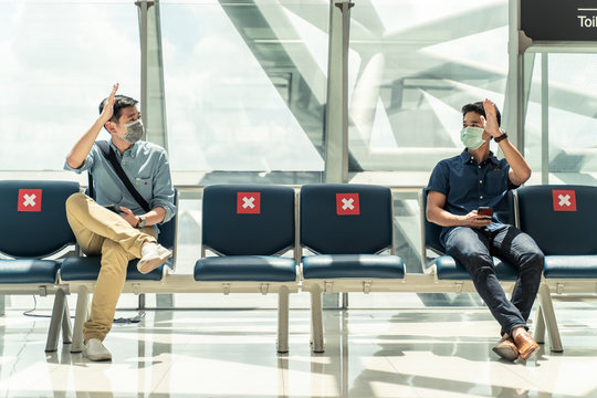 Social distancing, two men wearing face mask sitting keeping distance away from each other to prevent covid19 infection during pandemic. Empty chair seat red cross shows avoidance in airport terminal.