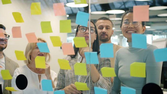 Multi-ethnic group of people is using colorful sticky notes on glassboard discussing business at work in modern office. Creative activity and teamwork concept.