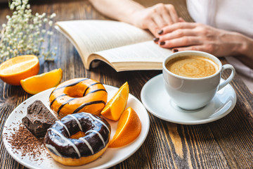 Woman is reading a book and having breakfast. Meal consists of cup of natural black coffee with fresh orange, donuts with orange and chocolate. Bright summer Breakfast, a tasty start to the morning