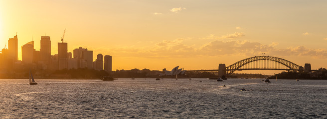 Obraz premium Panoramic view of Sydney downtown, harbor bridge and opera house with boats sailing in the bay at sunset. Beautiful orange sky.