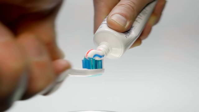 A man squeezes toothpaste from a tube onto a blue toothbrush on a white background. Closeup. The camera moves around