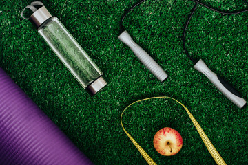 yoga mat, water bottle, jumping rope, measuring tape and an apple on green grass. Fitness life.