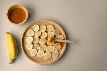 Top down view banana slices on light brown wooden plate with bowl of honey and honey dipper on plain grey color background.