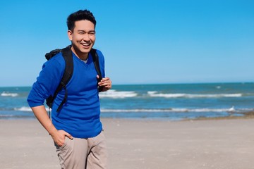 Happy Asian man backpacker in blue shirt standing by the beach.