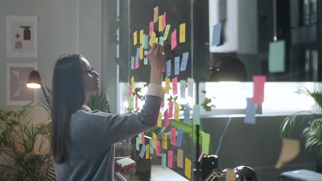 Young Business Woman Brainstorming. Asian Woman Writing Down Ideas On Sticky Notes Attached To Glass Wall. Business Success Concept