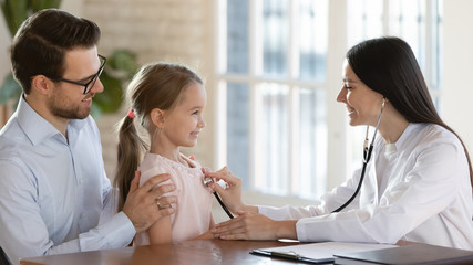 Fototapeta premium Young father with little preschooler daughter visit pediatrician for regular checkup examination at hospital, smiling female doctor GP listen to small girl child patient heartbeat at consultation