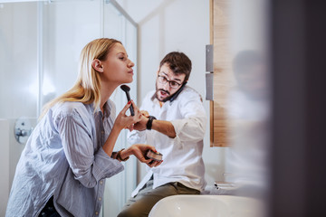 Handsome caucasian couple standing in bathroom and preparing for work. Man talking on the smart phone and looking at wristwatch while woman standing in front of mirror and putting powder.