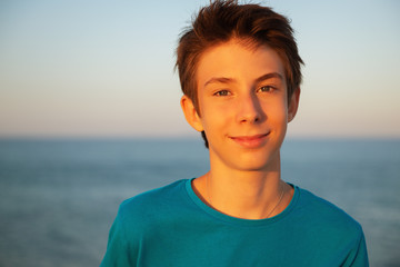 Handsome young boy enjoing life at beach at sunset. Beautiful tan smiling teen boy wearing blue t-shirt posing alone at Mediterranean sea coast. Travel, summer vacation, tourism, teenage lifestyle.