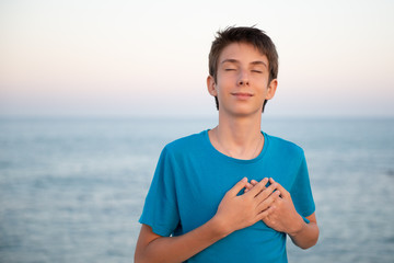Handsome young happy boy with eyes closed at beach holds hands on chest, sunrise or sunset....