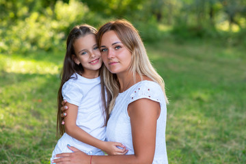 Fototapeta premium Little girl hugs mother, summer nature outdoor. Mom and daughter wearing white clothes against summer greenery. Family walks in park. Trust, kindness, maternity, parenthood, confidence, mother's love