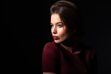 Fototapeta premium Portrait of attractive blue-eyed young woman over black background. Amazing girl with perfect makeup wearing elegant deep red dress. Beauty and fashion